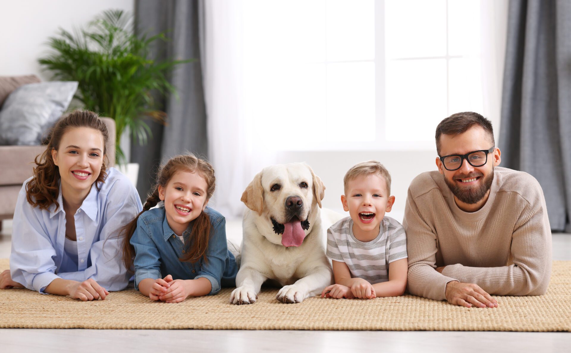 Smiling mother and father with little kids and adorable purebred Labrador retriever dog lying on floor and looking at camera while spending time together at home